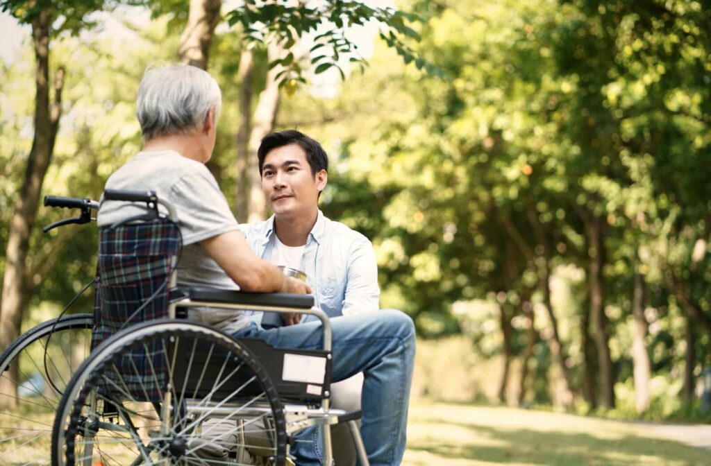 An adult kneels beside their senior parent while out for a walk in the park to talk to them bette