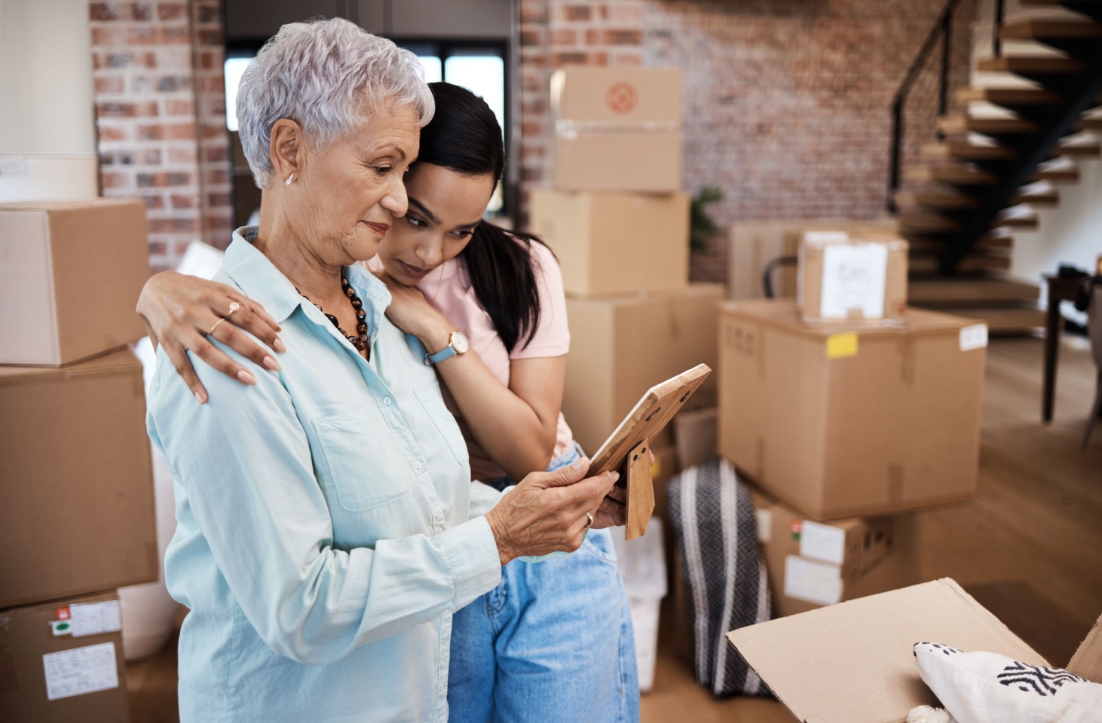 Older adult woman and younger family member embracing while looking at a photo, surrounded by moving boxes.