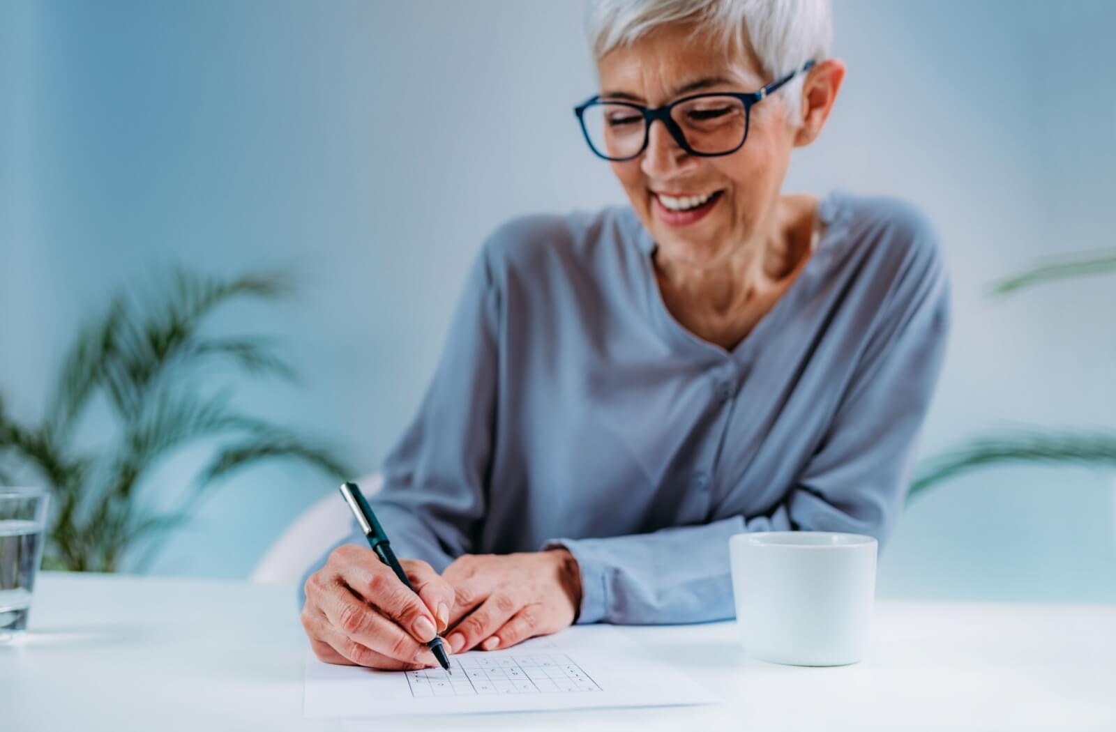 Smiling older woman doing sudoku on desk with white mug.