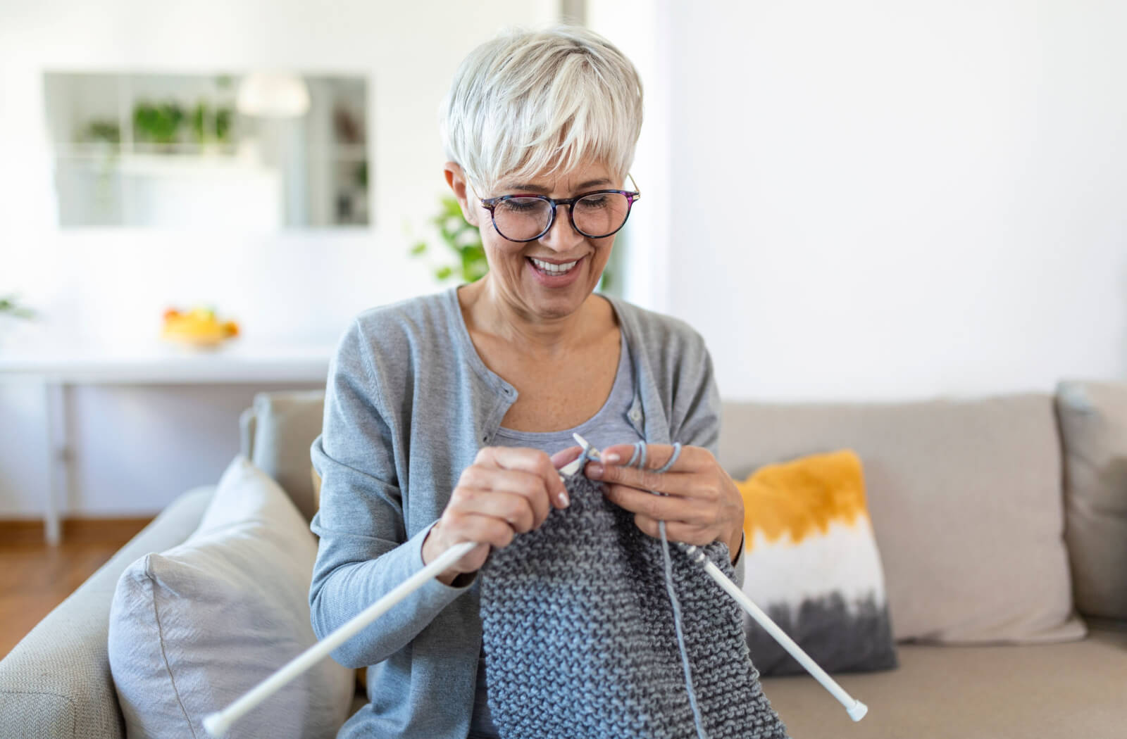An older adult knitting a scarf while sitting on their couch in a senior living community.