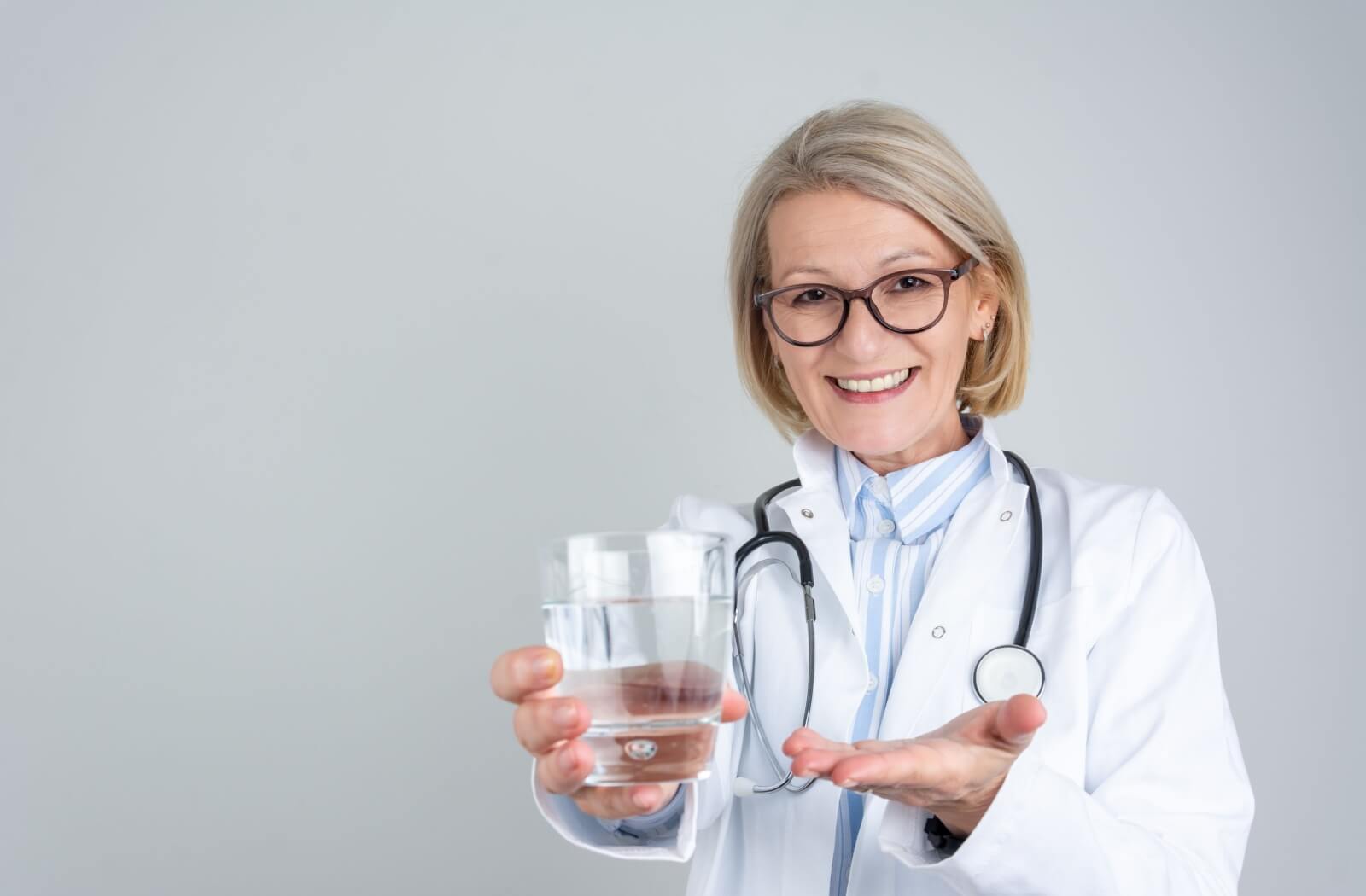 Against a grey backdrop, an older doctor smiles while holding out a glass of water to encourage hydration