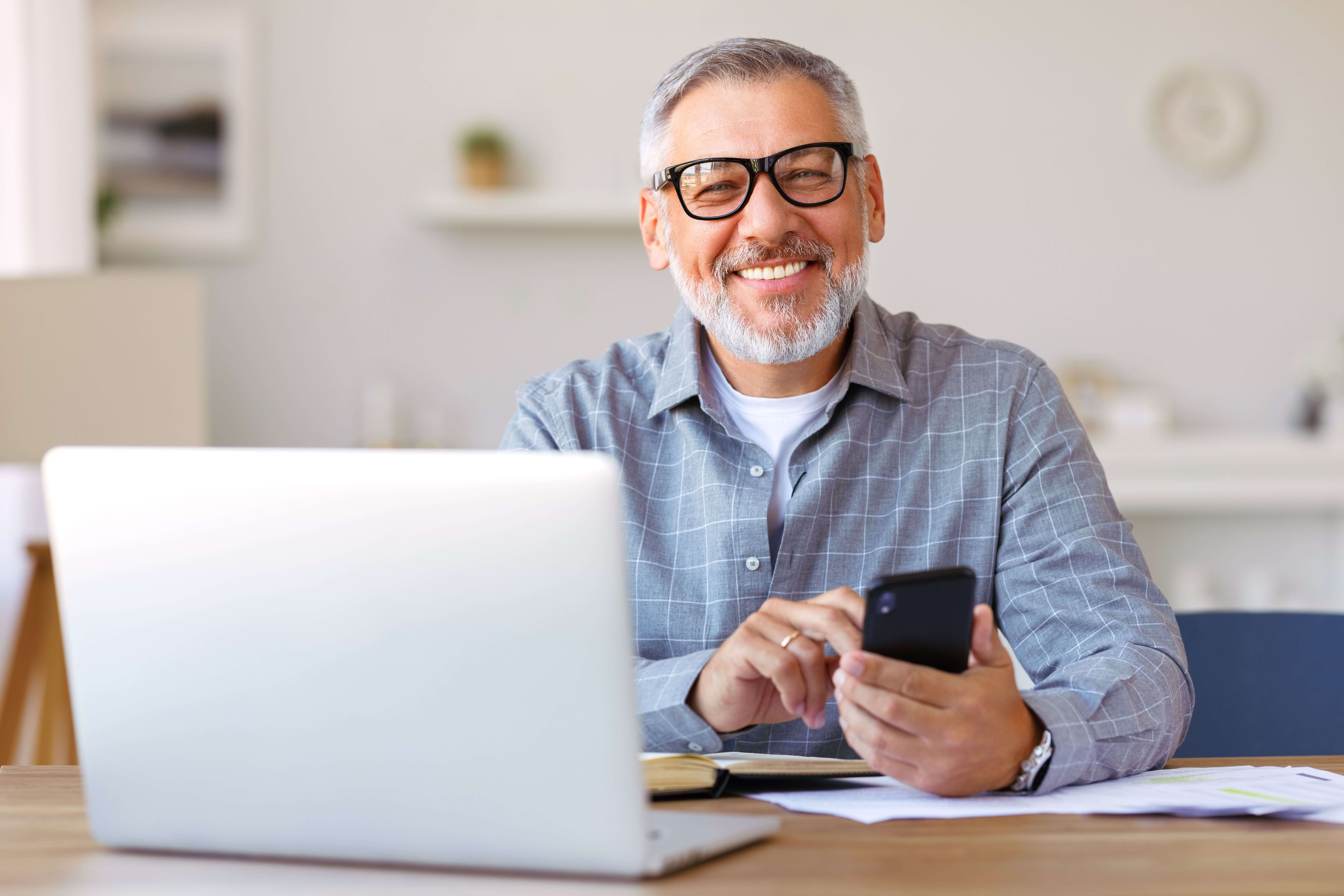 Senior smiling while holding phone, and laptop in front of them,