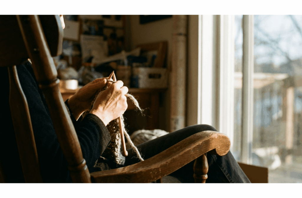 Adult sitting in a wooden rocking chair knitting with wool by a sunlit window, focusing on hands and yarn.